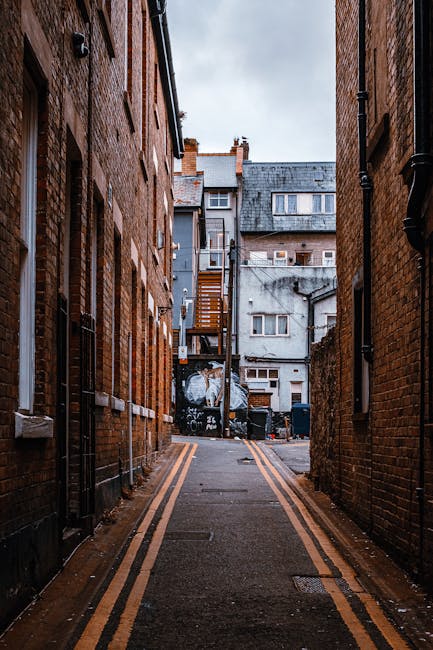 A narrow residential street in Queen's Park with a row of terraced houses featuring brick and white-painted facades, small front gardens with potted plants, and black metal railings. The pavement is c